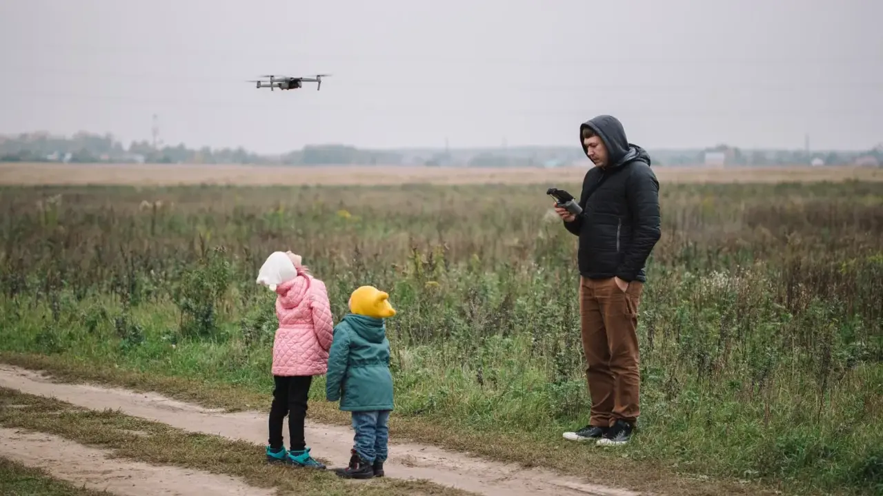 Ojciec steruje dronem, a dzieci z zaciekawieniem patrzą w niebo. Rodzinna przygoda z technologią w tle jesiennego krajobrazu.
