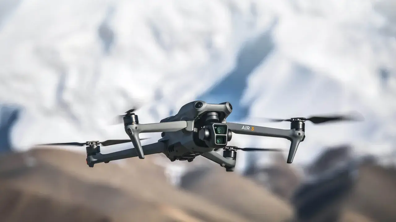 Szary dron DJI Air 3 z podwójną kamerą unosi się w powietrzu na tle rozmytych górskich szczytów.