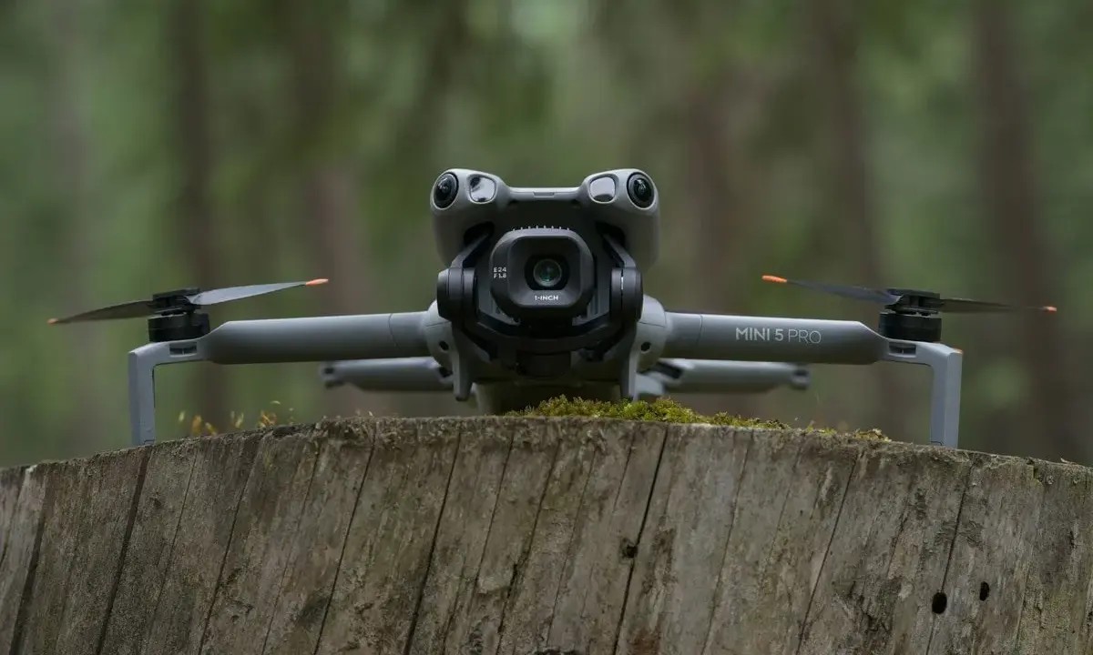 Szary dron DJI Mini 5 Pro z kamerą na gimbalu spoczywa na pniu drzewa w lesie.