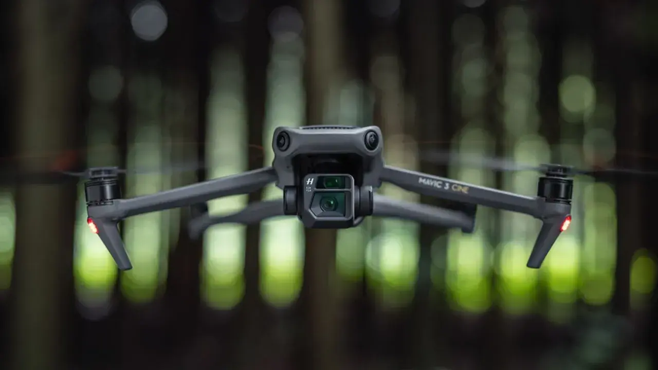 Szary dron Mavic 3 Cine zawieszony w powietrzu, z rozmytym zielonym lasem w tle.