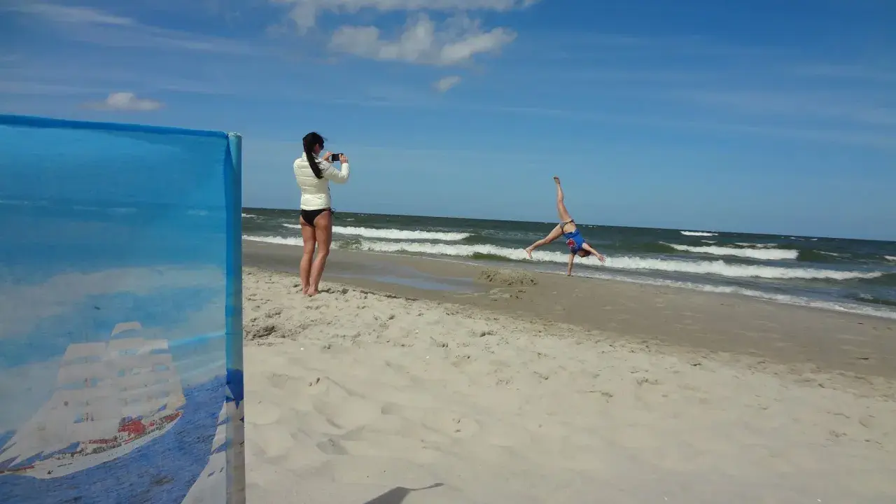Dziewczyna robi fikołka na plaży, podczas gdy druga filmuje ją telefonem. Krzywy horyzont dodaje dynamiki scenie.