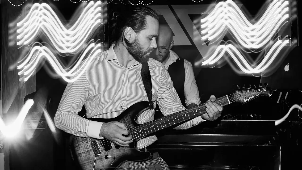 Muzyk gra na gitarze elektrycznej, a za nim stoi drugi mężczyzna. Efekt fal świetlnych sugeruje dynamiczną ekspozycję co to za moment.
