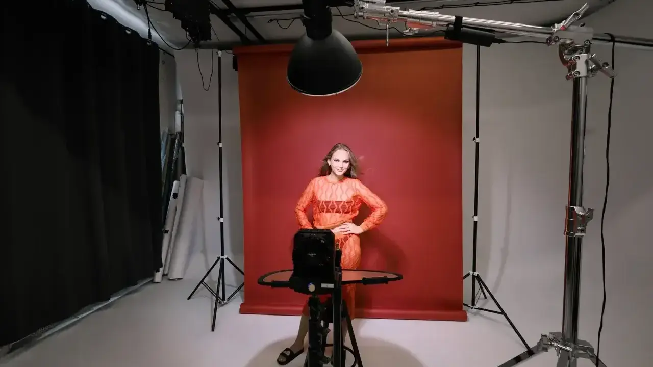 Oświetlenie studyjne przykłady: modelka w pomarańczowej sukience pozuje na tle czerwonego tła, otoczona sprzętem fotograficznym i lampami.
