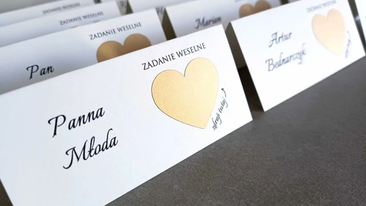 Białe karty z imionami gości i złotymi sercami, zapowiadające śmieszne zadania dla gości weselnych.