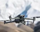 Szary dron DJI Air 3 z podwójną kamerą unosi się w powietrzu na tle rozmytych górskich szczytów.