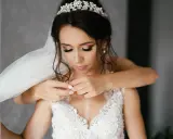 Panna młoda z welonem i diademem, jej świadkowa pomaga jej z naszyjnikiem.