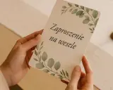 Dłonie trzymają zaproszenie na wesele z motywem liści. To ważny moment, gdy para młoda odpowiada na pytania o ślub.