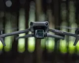 Szary dron Mavic 3 Cine zawieszony w powietrzu, z rozmytym zielonym lasem w tle.