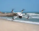 Biały dron DJI Lito X1 unosi się nad piaszczystą plażą, z morzem i niebem w tle.