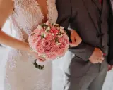 Panna młoda w koronkowej sukni trzyma bukiet różowych kwiatów, obok pan młody w garniturze. To piękny obrazek, jakby licencja na ślub była w ich rękach.