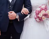 Młoda para, ona w sukni ślubnej, on w granatowym garniturze, trzymają się pod rękę. Bukiet róż w dłoni panny młodej.