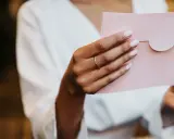 Dłoń z pierścionkiem trzyma różową kopertę. Zastanawiasz się, ile wcześnie zaprasza się na ślub?