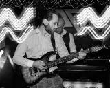 Muzyk gra na gitarze elektrycznej, a za nim stoi drugi mężczyzna. Efekt fal świetlnych sugeruje dynamiczną ekspozycję co to za moment.