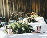 Eleganckie dekoracje stołu weselnego z geometrycznymi lampionami i zielenią. Informacja o noclegu dla gości weselnych tekst może być umieszczona obok.