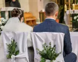 Para młoda siedzi w kościele. Krzesła ozdobione są zielonymi gałązkami. Kto może być świadkiem na ślubie?