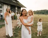 Rodzina w białych strojach, mama z córką na rękach, na tle wiejskiego domu. Skromny ślub kościelny bez wesela.