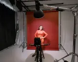 Oświetlenie studyjne przykłady: modelka w pomarańczowej sukience pozuje na tle czerwonego tła, otoczona sprzętem fotograficznym i lampami.