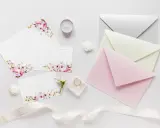 Różne rozmiary papieru i kopert w pastelowych kolorach, ozdobione kwiatami, wstążką i obrączkami.