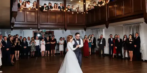 Para młoda tańczy przy dźwiękach wzruszających piosenek na ślub. Goście obserwują z balkonu i sali, niektórzy filmują telefonami.