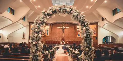 W kościele, pod kwiecistym łukiem, para młoda klęczy przed ołtarzem. Wokół goście, świadkowie, ksiądz. Piękna sceneria ślubna, pełna nadziei i tradycji, jak przesądy ślubne.