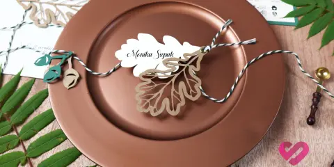 Winietki co to? Eleganckie winietki z imieniem "Monika Szpak" na liściu dębu, ozdobione sznurkiem i żołędziami, na brązowym talerzu.