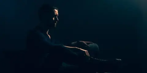 Mężczyzna w ciemnym pomieszczeniu, siedzi w fotelu. Jego sylwetka jest ledwo widoczna, podkreślona przez subtelne światło. To idealny przykład low key fotografii.