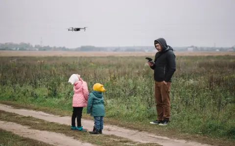 Ojciec steruje dronem, a dzieci z zaciekawieniem patrzą w niebo. Rodzinna przygoda z technologią w tle jesiennego krajobrazu.