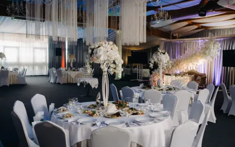 Elegancka sala weselna z białymi obrusami, kwiatami i oświetleniem tworząca magiczną atmosferę. To idealne miejsce na niezapomniane atrakcje na wesele.