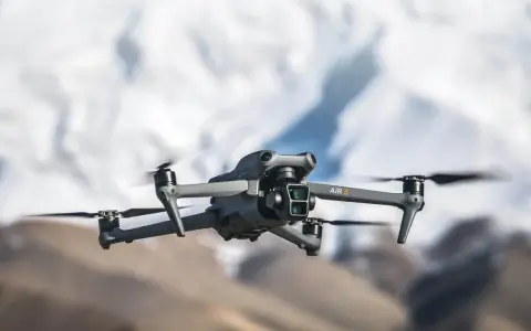 Szary dron DJI Air 3 z podwójną kamerą unosi się w powietrzu na tle rozmytych górskich szczytów.