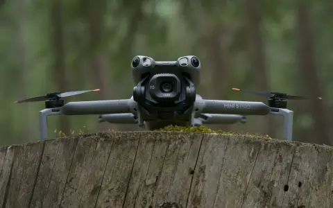Szary dron DJI Mini 5 Pro z kamerą na gimbalu spoczywa na pniu drzewa w lesie.