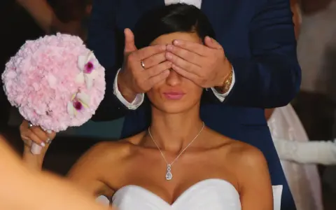 Pan młody zasłania oczy pannie młodej, która trzyma bukiet. To tradycja oczepin, co to za zwyczaj?
