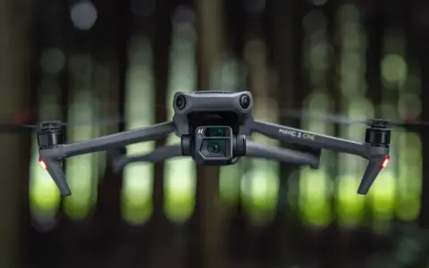 Szary dron Mavic 3 Cine zawieszony w powietrzu, z rozmytym zielonym lasem w tle.