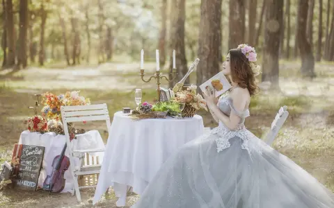 Kobieta w sukni ślubnej czyta książkę przy stole zastawionym owocami i szampanem. Sceneria leśna, atmosfera spokoju przed weselem.