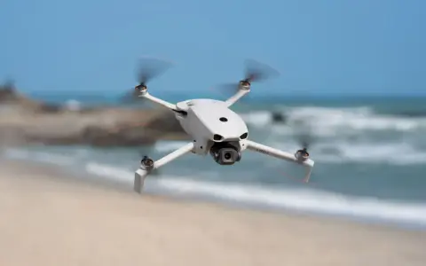 Biały dron DJI Lito X1 unosi się nad piaszczystą plażą, z morzem i niebem w tle.