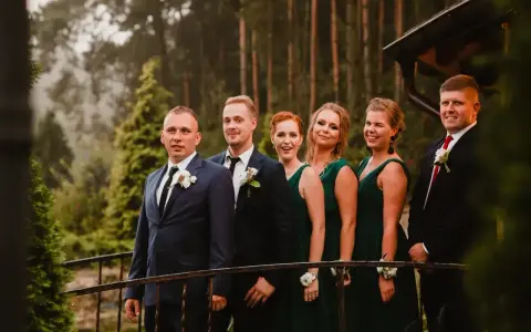 Pan młody i jego drużba, ubrani w garnitury, stoją obok druhen w zielonych sukniach. To idealna drużyna, która wie, co potrzebuje świadek do ślubu kościelnego.