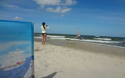 Dziewczyna robi fikołka na plaży, podczas gdy druga filmuje ją telefonem. Krzywy horyzont dodaje dynamiki scenie.