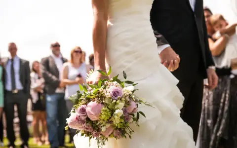 Panna młoda z bukietem, trzymająca pana młodego za rękę. W tle goście weselni. Czy osoba towarzysząca płaci za wesele?