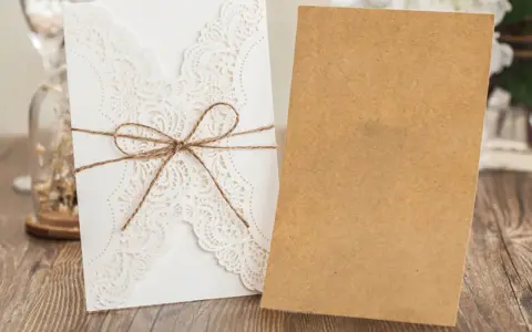 Eleganckie, białe zaproszenie ślubne z koronkowym wzorem, przewiązane sznurkiem. Obok leży brązowa koperta, gotowa na ręczne wypisywanie zaproszeń ślubnych.