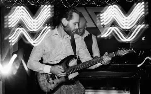 Muzyk gra na gitarze elektrycznej, a za nim stoi drugi mężczyzna. Efekt fal świetlnych sugeruje dynamiczną ekspozycję co to za moment.