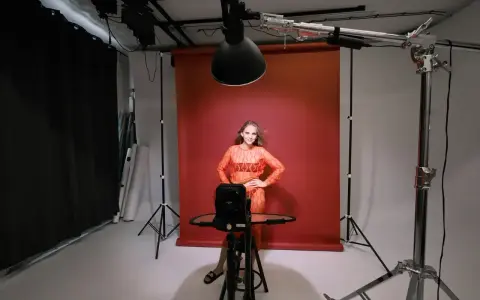 Oświetlenie studyjne przykłady: modelka w pomarańczowej sukience pozuje na tle czerwonego tła, otoczona sprzętem fotograficznym i lampami.