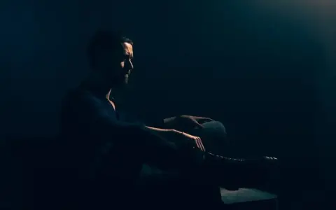 Mężczyzna w ciemnym pomieszczeniu, siedzi w fotelu. Jego sylwetka jest ledwo widoczna, podkreślona przez subtelne światło. To idealny przykład low key fotografii.