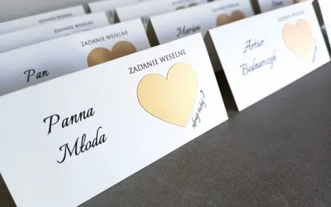 Białe karty z imionami gości i złotymi sercami, zapowiadające śmieszne zadania dla gości weselnych.