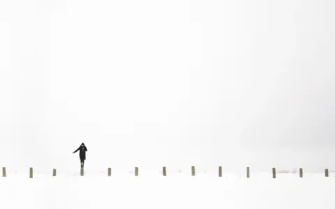 Osoba w czarnym płaszczu balansuje na słupku w śnieżnym krajobrazie. Minimalistyczna, high key fotografia.