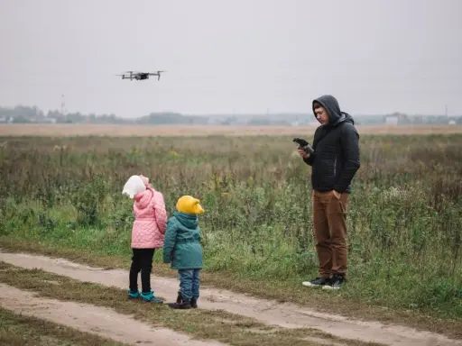 Ojciec steruje dronem, a dzieci z zaciekawieniem patrzą w niebo. Rodzinna przygoda z technologią w tle jesiennego krajobrazu.