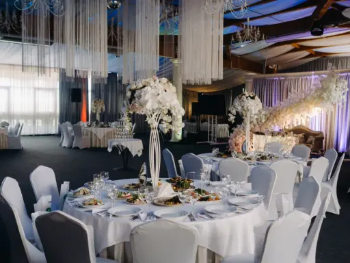 Elegancka sala weselna z białymi obrusami, kwiatami i oświetleniem tworząca magiczną atmosferę. To idealne miejsce na niezapomniane atrakcje na wesele.