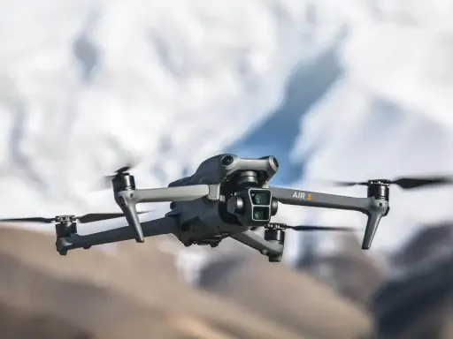 Szary dron DJI Air 3 z podwójną kamerą unosi się w powietrzu na tle rozmytych górskich szczytów.