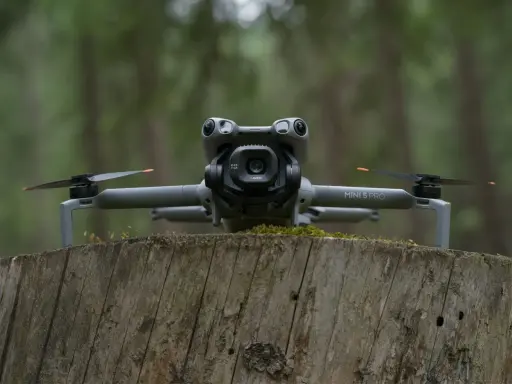 Szary dron DJI Mini 5 Pro z kamerą na gimbalu spoczywa na pniu drzewa w lesie.