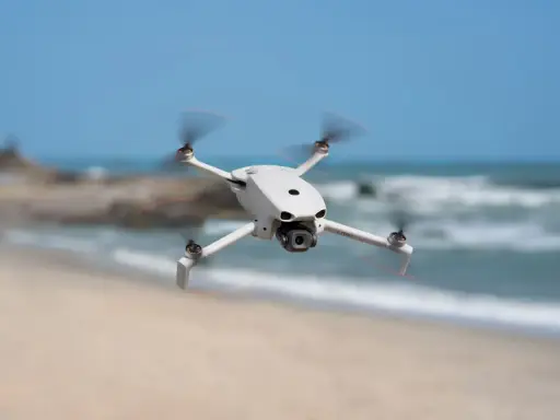 Biały dron DJI Lito X1 unosi się nad piaszczystą plażą, z morzem i niebem w tle.