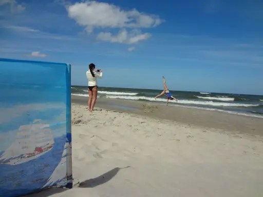 Dziewczyna robi fikołka na plaży, podczas gdy druga filmuje ją telefonem. Krzywy horyzont dodaje dynamiki scenie.