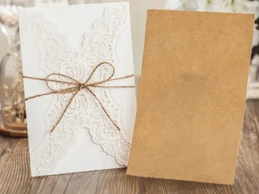 Eleganckie, białe zaproszenie ślubne z koronkowym wzorem, przewiązane sznurkiem. Obok leży brązowa koperta, gotowa na ręczne wypisywanie zaproszeń ślubnych.
